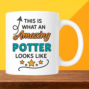 May include: White ceramic mug with the text "THIS IS WHAT AN Amazing POTTER LOOKS LIKE" in black, orange, and teal. The mug has a curved handle and is decorated with star graphics.
