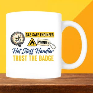 May include: White ceramic mug with a humorous design. The mug features text that reads "GAS SAFE ENGINEER", "Hot Stuff Handler", and "TRUST THE BADGE". The design includes a gauge, a flame symbol, and a permit tag.