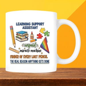 May include: White ceramic mug with a colourful design featuring the text "LEARNING SUPPORT ASSISTANT" and other phrases. The design includes a pencil, books, glue, puzzle pieces, and a rainbow. The mug is on a wooden surface.