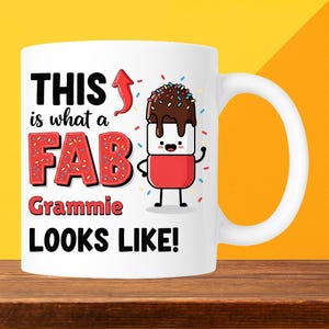 May include: White ceramic mug with a handle. The mug features the text "THIS is what a FAB Grammie LOOKS LIKE!" in black and red lettering. A cartoon marshmallow character with chocolate and sprinkles is also on the mug.