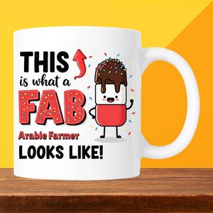 May include: White ceramic mug with a fun design. The mug displays the text "THIS is what a FAB Arable Farmer LOOKS LIKE!" and a cartoon marshmallow character with chocolate and sprinkles. The mug has a white handle.