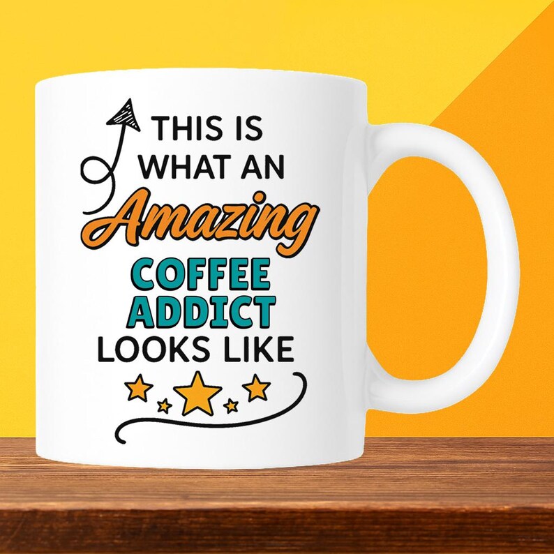 Op de afbeelding: Witte keramische koffiemok met de tekst "THIS IS WHAT AN Amazing COFFEE ADDICT LOOKS LIKE" in zwart, oranje en teal. De mok heeft een gebogen handvat en staat tegen een gele en oranje achtergrond.
