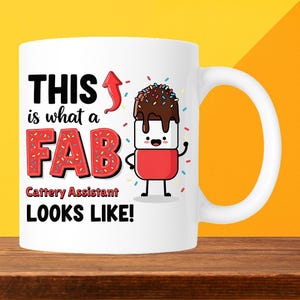 May include: White ceramic mug with the text "THIS is what a FAB Cattery Assistant LOOKS LIKE!" and a cartoon marshmallow character with chocolate frosting. The mug has a red arrow and a red and pink "FAB" design.