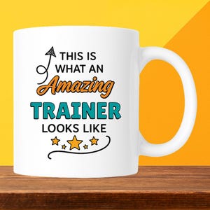 May include: White ceramic mug with the text "THIS IS WHAT AN Amazing TRAINER LOOKS LIKE" in black, orange, and teal. The mug has a curved handle and is set against a yellow and orange background.