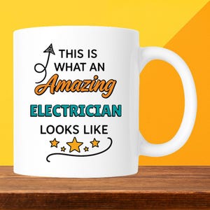 May include: White ceramic mug with the text "THIS IS WHAT AN Amazing ELECTRICIAN LOOKS LIKE" in black, orange, and teal. The mug has a curved handle and is set against a yellow and orange background.