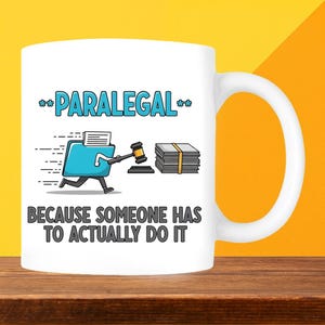 May include: White ceramic mug with a humorous design for paralegals. The mug features the word "PARALEGAL" above a cartoon image of a file folder holding a gavel and a stack of papers, with the text "BECAUSE SOMEONE HAS TO ACTUALLY DO IT" below.