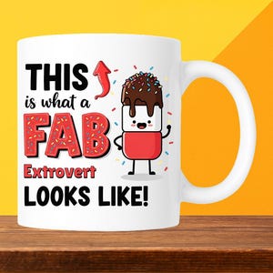 May include: White ceramic mug with a handle. The mug has the text "THIS is what a FAB Extrovert LOOKS LIKE!" in black and red. A cartoon marshmallow character with chocolate frosting and sprinkles is also on the mug.