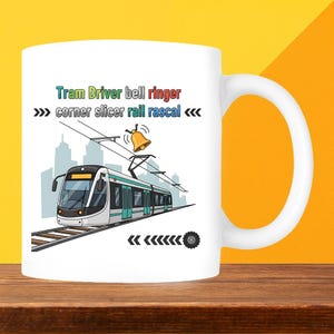 May include: White ceramic mug with a tram design and the text "Tram Driver bell ringer corner slicer rail rascal". The tram is white and teal, with a cityscape background and a bell icon.