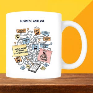 May include: White ceramic mug featuring the words "BUSINESS ANALYST" and a humorous graphic. The graphic includes speech bubbles with phrases like "SYNERGY" and "PARADIGM SHIFT".