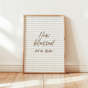 Op de afbeelding: Een ingelijste print met een licht houten frame. De print heeft een gestreepte achtergrond in grijstinten en wit. De woorden "How blessed are we" zijn geschreven in een cursief lettertype in een warme bruine kleur. De print wordt weergegeven op een houten vloer.