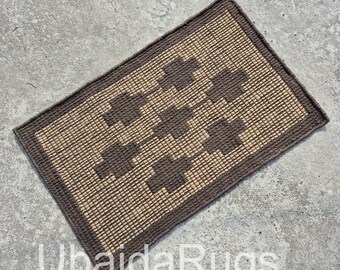 May include: Rectangular woven rug with a geometric pattern. The rug features a light brown center with dark brown cross-shaped designs and a dark brown border. The text "UbaidaRugs" is at the bottom.
