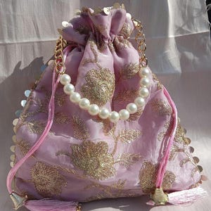 May include: A lavender drawstring bag with gold floral embroidery and a pearl handle. The bag features gold sequins around the top edge, a gold chain handle, and pink tassels. The bag is made of a soft, silky material.