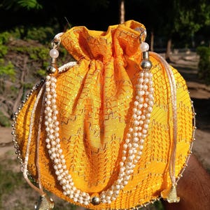 May include: A round, yellow pouch with a drawstring closure. The pouch is decorated with a beaded strap and gold accents. The fabric has a textured pattern. The pouch is designed to hold small items.