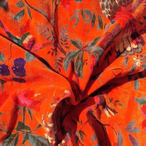 May include: Close-up of a vibrant orange fabric with a floral pattern. The design features red, pink, purple, and blue flowers, along with green and grey leaves and branches. The fabric has a soft, velvety texture.