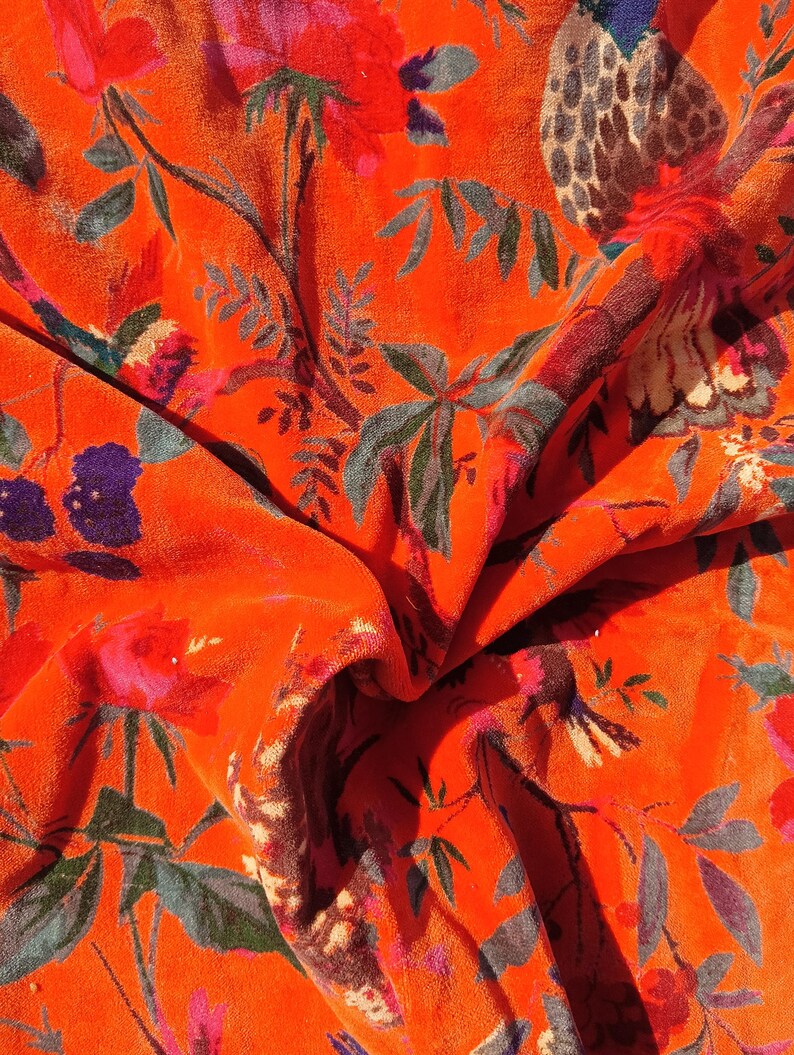 May include: Close-up of a vibrant orange fabric with a floral pattern. The design features red, pink, purple, and blue flowers, along with green and grey leaves and branches. The fabric has a soft, velvety texture.