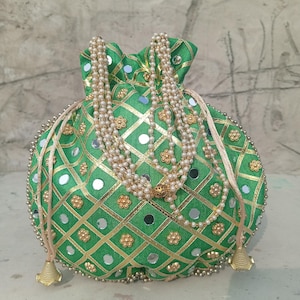 Op de afbeelding: Groene trekkoordtas met gouden en zilveren versieringen. De tas heeft een geometrisch patroon met gouden randen, kleine spiegels en gouden bloemaccenten. Een parelketting drapeert over de bovenkant. De tas heeft gouden kwastjes.