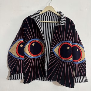 May include: Black zip-up jacket with embroidered eye designs. The jacket features large, stylized eyes with red, blue, and gold accents radiating pink lines. The interior lining and collar have a black and white striped pattern.