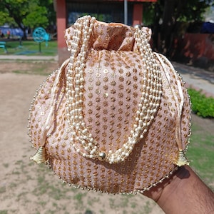 May include: A blush pink drawstring pouch with gold floral embroidery and a beaded pearl strap. The bag has gold tassels and a beaded trim. The pouch is held up by a hand.