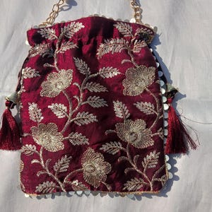 Może przedstawiać: Bordowa torebka ze sznurkiem ze złotym haftem kwiatowym i obszyciem z cekinów. Torebka ma uchwyt z pereł i złote łańcuszki. Bordowe frędzle zwisają z boków. Torebka jest przeznaczona do przenoszenia drobnych przedmiotów.
