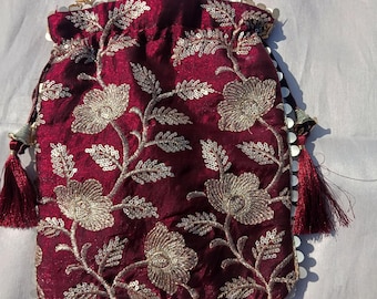 May include: Burgundy drawstring bag with gold floral embroidery and sequin trim. The bag features a pearl beaded handle and gold chain accents. Burgundy tassels hang from the sides, adding a decorative touch. The bag is likely used for carrying small items.