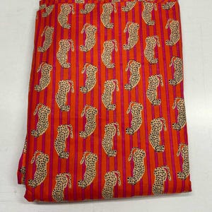 May include: Folded fabric with a repeating pattern of tan leopard-like figures on a bright orange background with vertical pink stripes. The fabric appears to be cotton or a similar material, suitable for various sewing projects.