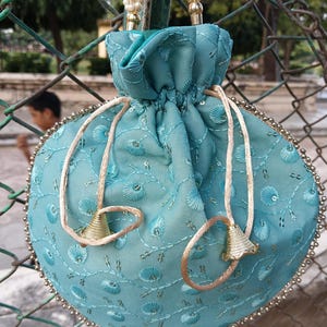 May include: A teal drawstring pouch with detailed floral embroidery and a beaded border. The bag has a drawstring closure with gold-coloured bell-shaped accents and a pearl-beaded handle. The pouch is hanging on a chain-link fence.