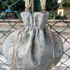 Può includere: Una borsa a coulisse grigio chiaro con ricami floreali e paillettes dorate. La borsa ha un manico di perline e nappe dorate. La borsa rotonda ha un bordo di perline sul bordo.