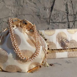 May include: Two elegant, off-white fabric bags with gold circular patterns. One is a drawstring pouch with a beaded handle, the other a rectangular clutch with a decorative clasp and fringe. Both bags have gold accents.
