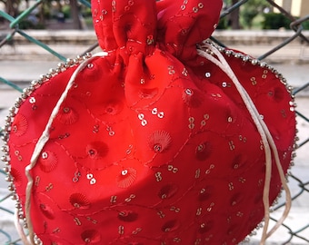 May include: A red drawstring pouch with gold embellishments and a beaded handle. The pouch is decorated with sequins and floral embroidery. The handle is made of white beads and gold accents. The pouch has a gold tassel at the end of the drawstring.