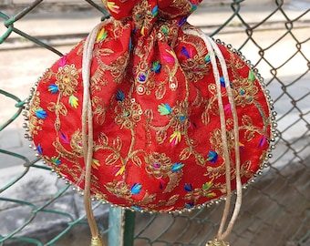 Könnte beinhalten: Ein roter, runder Beutel mit Kordelzug, aufwendiger Goldstickerei und farbenfrohen Akzenten. Die Tasche hat einen Perlenbesatz und goldene Quasten an den Kordelzügen. Die Tasche hängt an einem Maschendrahtzaun.