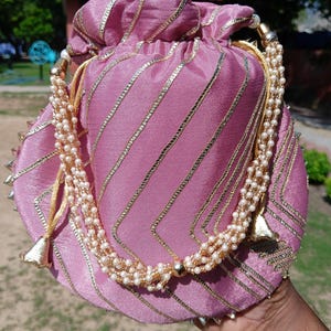 May include: A pink satin drawstring bag with gold and silver detailing. The bag has diagonal gold stripes and a beaded strap with a gold tassel. The bag is round and has a decorative design.