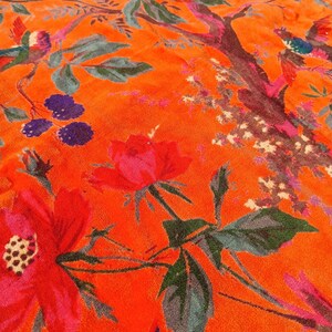May include: Close-up of a vibrant orange fabric with a floral and bird pattern. The design features large red flowers, green leaves, and colourful birds perched on branches. The fabric has a soft, textured appearance.