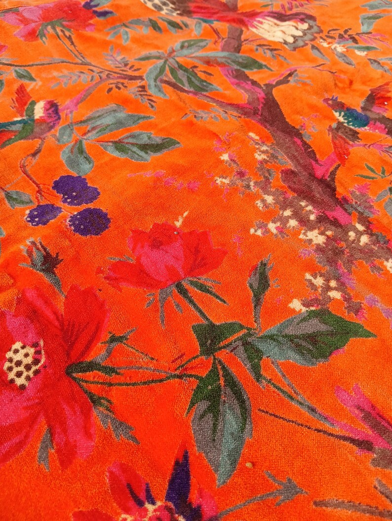 May include: Close-up of a vibrant orange fabric with a floral and bird pattern. The design features large red flowers, green leaves, and colourful birds perched on branches. The fabric has a soft, textured appearance.