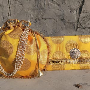 Könnte beinhalten: Zwei gelbe Stoffhandtaschen. Eine ist eine Kordelzugtasche mit einem perlenbesetzten Riemen und goldenen Akzenten. Die andere ist eine rechteckige Clutch mit einem dekorativen silbernen Verschluss und goldenen Verzierungen. Beide haben ein goldenes kreisförmiges Muster.
