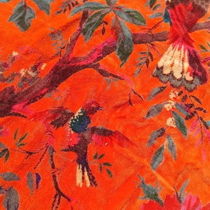 May include: Bright orange fabric with a detailed floral and bird pattern. The design features colourful birds with red, blue, and white feathers, set against a backdrop of green leaves and red flowers. The fabric has a soft, textured appearance.