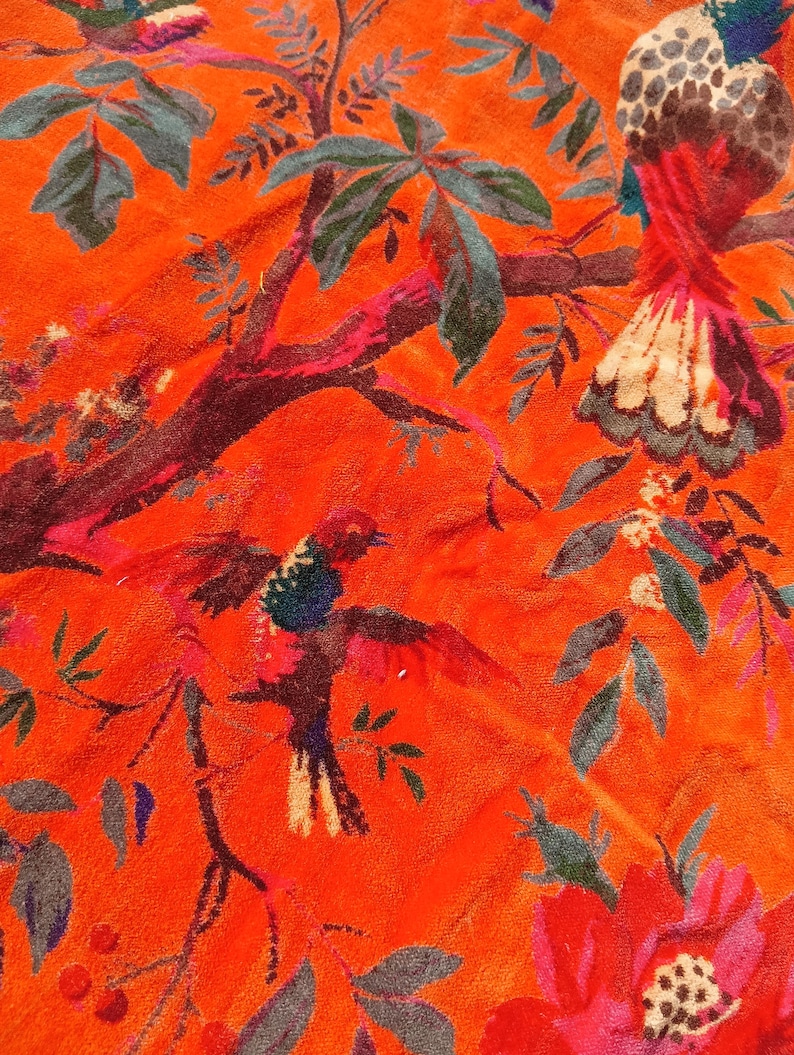 May include: Bright orange fabric with a detailed floral and bird pattern. The design features colourful birds with red, blue, and white feathers, set against a backdrop of green leaves and red flowers. The fabric has a soft, textured appearance.