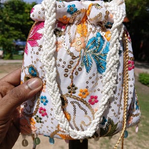 May include: White drawstring bag with colourful floral embroidery, gold sequins, and a braided white strap. The bag has a beaded drawstring and dangling beaded accents. The bag is held up by a metal pole.
