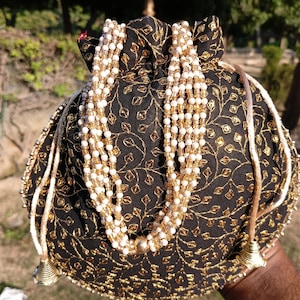 May include: Black drawstring pouch with gold floral embroidery and beaded accents. The pouch features a beaded necklace-like design and gold tassels. The bag is held by a person.
