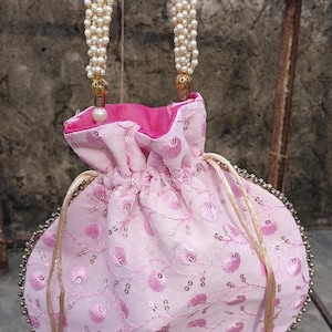 May include: A pink embroidered drawstring bag with a beaded edge and pearl-adorned handles. The bag features a floral pattern and a contrasting pink interior. Gold-coloured tassels hang from the bottom, adding a touch of elegance to this accessory.