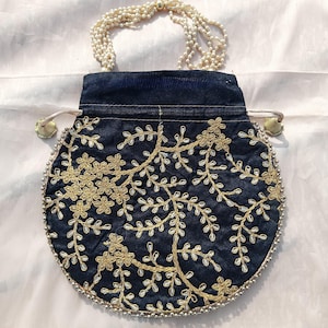 Op de afbeelding: Een marineblauwe, ronde handtas met gouden bloemenborduurwerk. De tas heeft een kralenhandvat en een trekkoordsluiting met gouden kwastjes. De randen zijn afgewerkt met kleine zilveren kralen. Geschikt voor speciale gelegenheden.