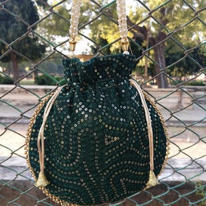 May include: A dark green drawstring bag with gold sequins and a beaded handle. The bag has a round shape with a gold beaded trim and gold tassels. The handle is made of small white beads.