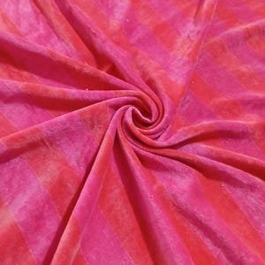 May include: Close-up of a soft, plush fabric with a striped pattern. The fabric features alternating bands of bright pink and red-pink hues. The material is gathered and swirled, creating a textured effect.
