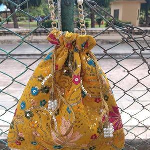 May include: Mustard yellow drawstring bag with detailed floral embroidery in pink, blue, and green. It has a pearl beaded handle and gold chain detailing. The base is trimmed with small, colourful beads.