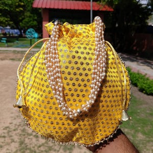 May include: A yellow drawstring bag with a gold floral pattern and a beaded pearl strap. The bag has gold trim and small bell-shaped accents. The bag is held in front of a blurred outdoor background.