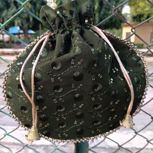 May include: Dark green drawstring pouch with gold sequins and a beaded trim. The pouch has a drawstring closure with gold-colored metal accents and a beaded handle. The bag is made of a textured fabric.