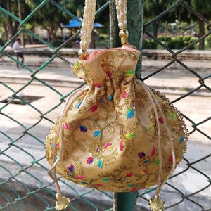 May include: A gold drawstring bag with detailed embroidery and colourful floral patterns. The bag has a beaded strap and decorative tassels. The bag is suspended from a green metal pole.