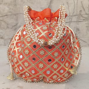 May include: An orange drawstring pouch with a gold geometric pattern, small mirrors, and floral accents. The pouch is adorned with a pearl necklace and gold tassels. The bag is a decorative accessory.