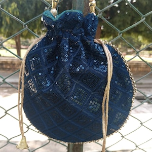 May include: A dark teal drawstring bag with a beaded handle and gold detailing. The bag features a geometric pattern and is embellished with sequins. The drawstring cords end in small, gold-coloured tassels.