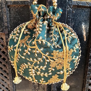 Op de afbeelding: Een ronde, donkerblauwe tas met trekkoord en gouden en witte bloemborduurwerk. De tas heeft een kralen hengsel en gouden kwastjes. De randen zijn afgewerkt met kleine, zilveren kralen. De tas is een decoratief accessoire.
