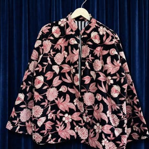 以下が含まれることがあります： 木製のハンガーに掛けられた、ピンクの花柄の黒いジャケット。 ジャケットはフルレングスのジッパーとスタンドカラーが付いています。 裏地はストライプです。 花柄のデザインには、さまざまな花と葉が含まれています。